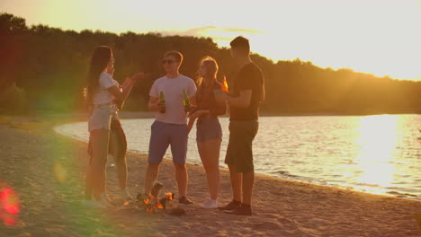 The-five-students-spend-time-at-sunset-on-the-sand-beach-in-shorts-and-t-shirts-around-bonfire-with-beer.-They-are-talking-to-each-other-and-enjoying-the-warm-summer-evening-near-the-river.